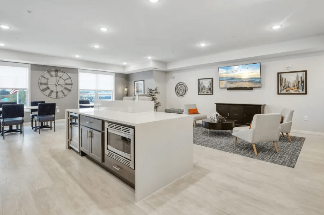 A modern living room and kitchen area with light wood floors, a large island counter, and built-in stainless steel appliances. The living room features a gray rug, white and gray armchairs, and a wall-mounted TV above a dark wooden cabinet. Large windows allow ample natural light.