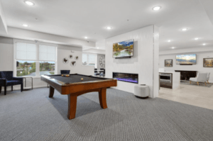 A modern recreation room featuring a billiards table with a cue stick and balls. The room has carpeted flooring, a large window with blinds, framed art on white walls, a wall-mounted TV above an electric fireplace, and various seating options in the background.