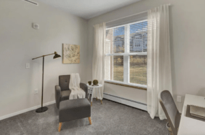 A cozy, sunlit corner of a room with a modern gray armchair, a matching ottoman, and a white side table. The armchair has a beige throw draped over it, and the table holds a small potted plant. A tall floor lamp stands beside the chair. Nearby, a desk and chair are partly visible. A large window with sheer white curtains lets in natural light.