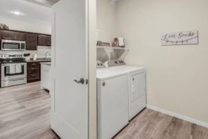 A modern laundry area with a washer and dryer side by side on the right. Above them is a shelf with a laundry basket. The adjacent kitchen features stainless steel appliances and dark wooden cabinets. A "Lost Socks" sign is mounted on the laundry room wall.
