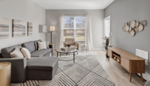 A modern living room with light grey walls, featuring a grey sectional sofa adorned with beige and white pillows, a round coffee table, a large window with sheer curtains, an armchair, a patterned rug, and a wooden TV stand with decorative items on the wall.
