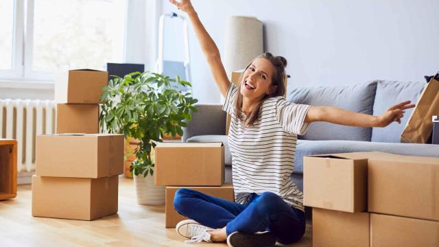 Woman sitting on the floor in a living room, surrounded by moving boxes. She is smiling and stretching her arms joyfully. A potted plant and a sofa are in the background.